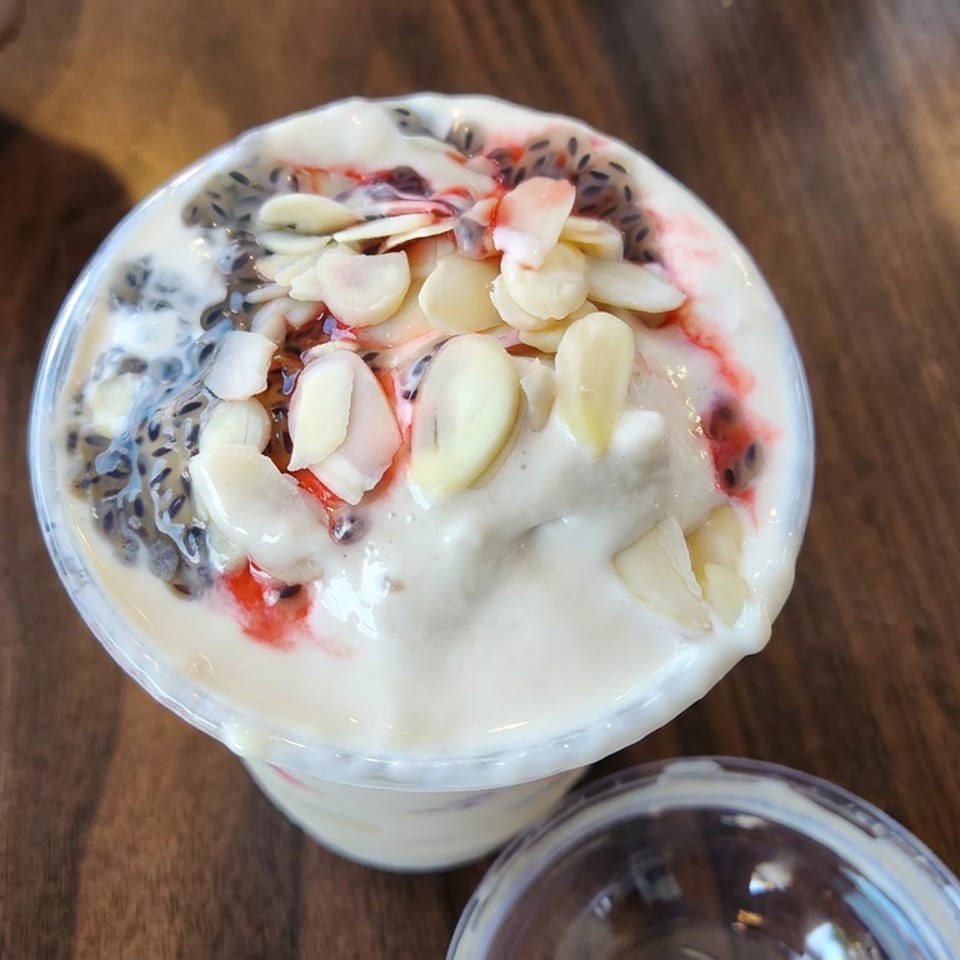 Creamy dessert topped with sliced almonds, red fruit sauce, and dark seeds in a clear cup on a wooden table.