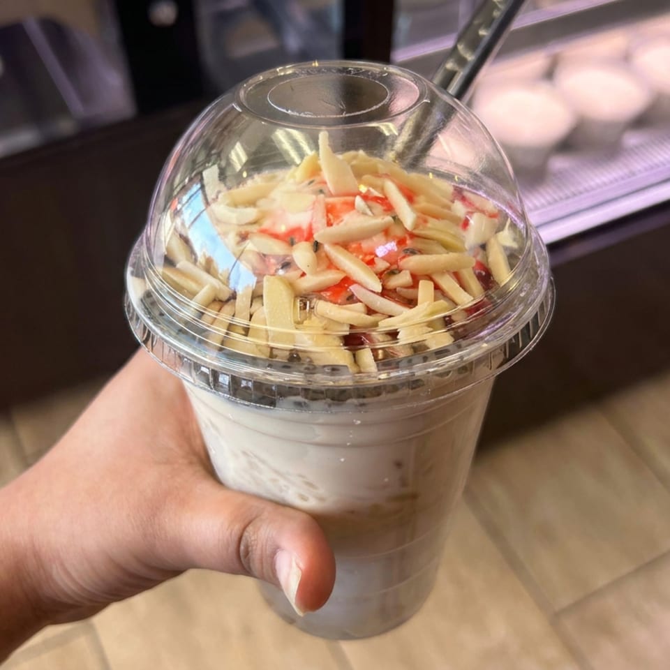 Hand holding a clear cup of creamy dessert topped with pale crunchy pieces and red fruit sauce at a restaurant counter.