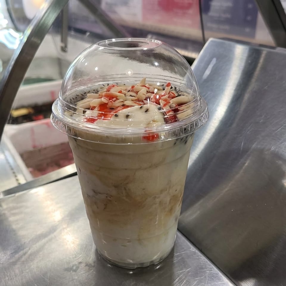 Layered dessert parfait topped with whipped cream, chopped nuts, and red fruit in a clear cup on a metal counter.
