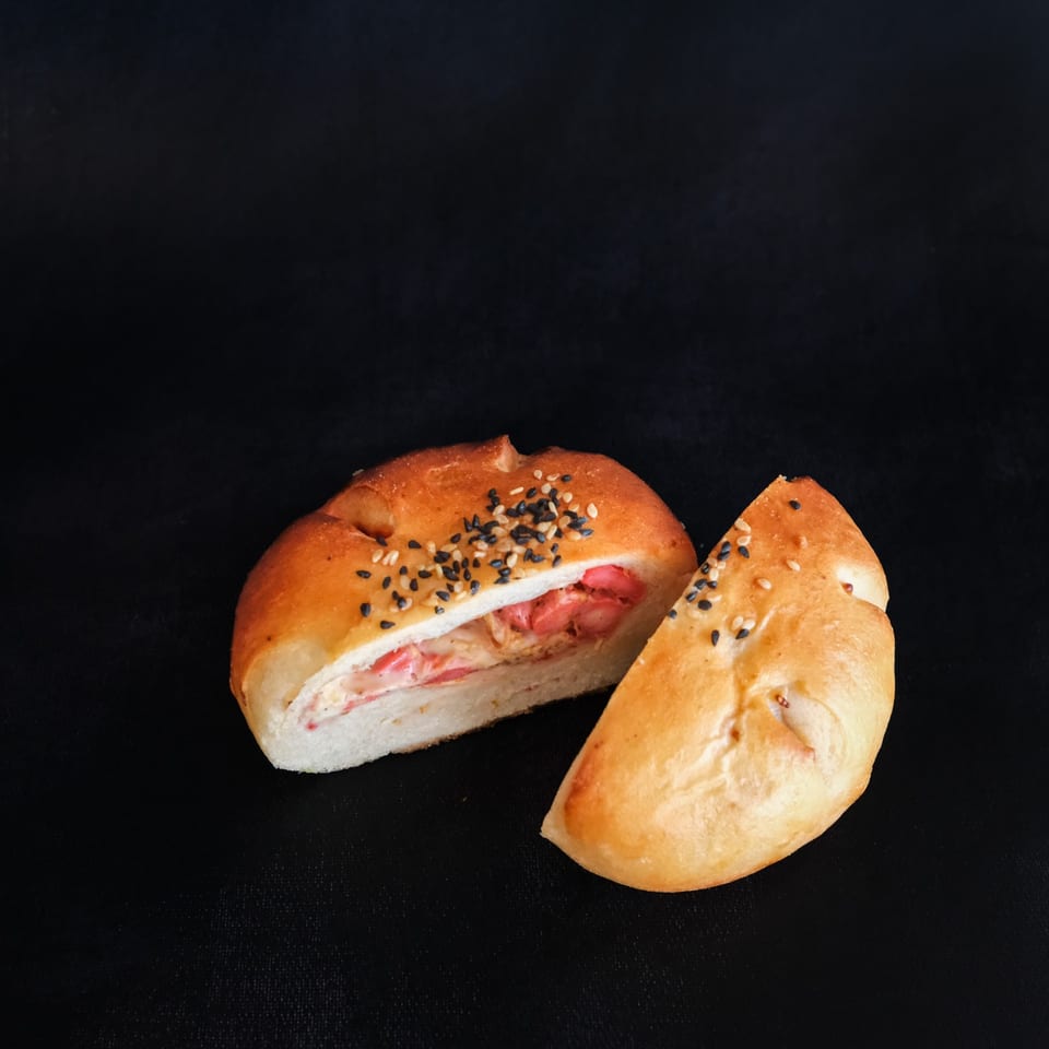 Sesame-topped stuffed bread rolls, one sliced open to reveal a savory pink and creamy filling, arranged on a black background.