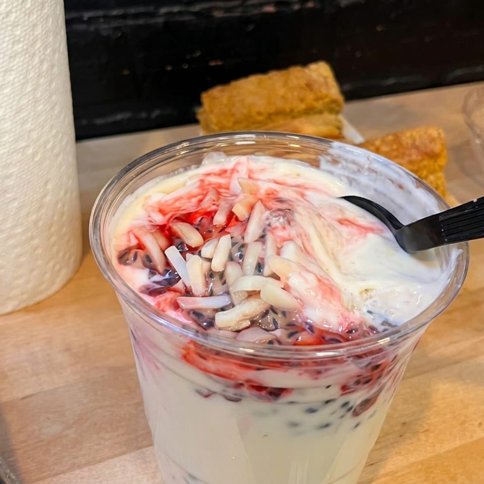 Clear cup of creamy yogurt layered with red fruit, dark berries, and shredded coconut on a wooden restaurant counter.