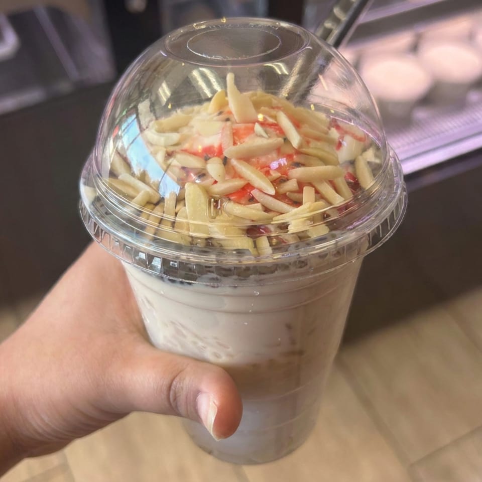 Hand holds a clear takeaway cup layered with creamy yogurt, fruit, and crunchy almond-like toppings inside a café.